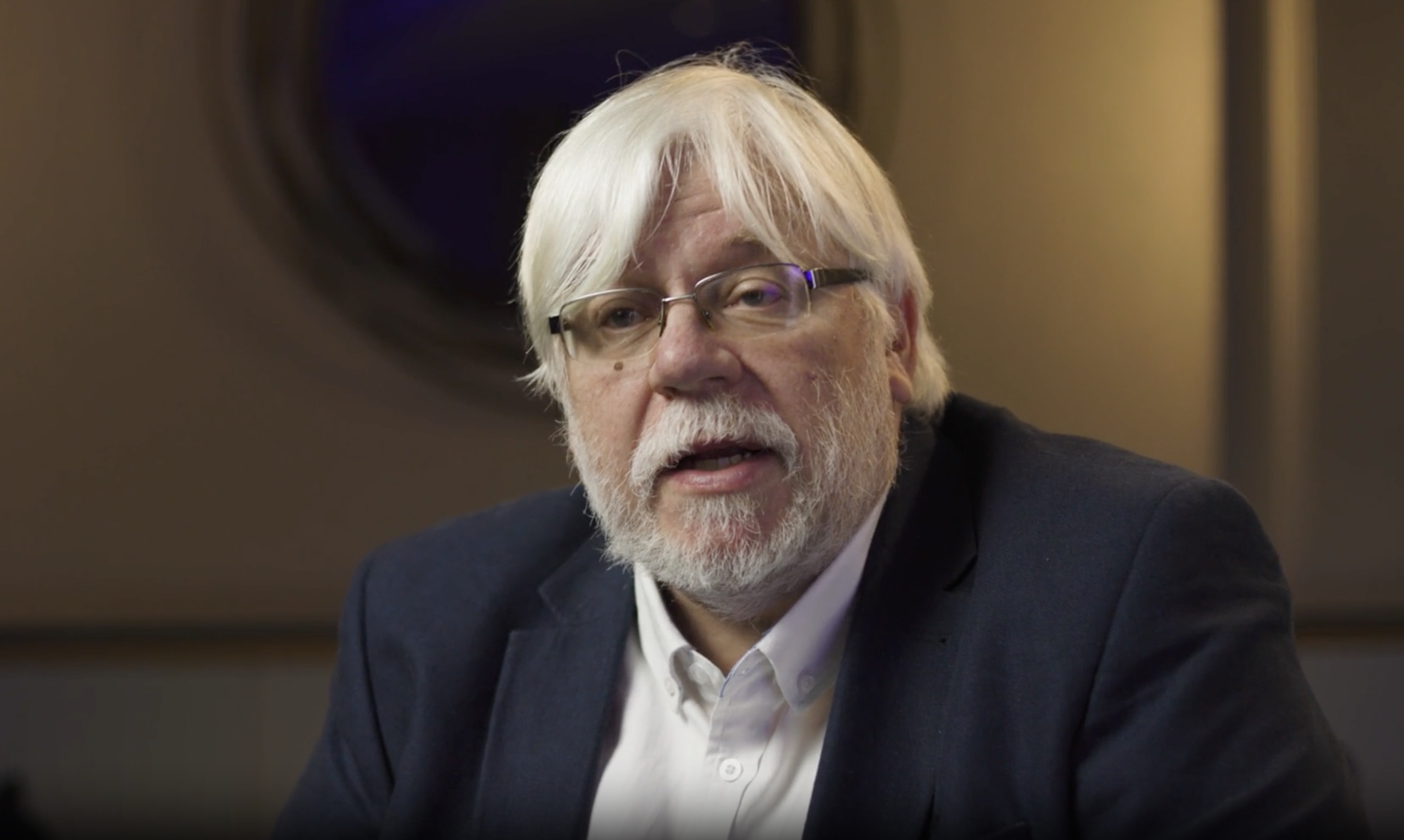An older man with white hair and a beard, wearing glasses, a dark suit, and a white shirt, speaks while seated indoors.